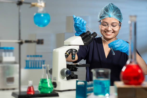 Young good looking Asian Indian Female Lab Technician wearing a navy blue uniform, protective eyeglasses, blue lab gloves, hairs nicely clipped behind with blue bouffant cap, light pastel orange lipstick, working on the microscope in the scientific research lab of a hospital, adding liquid using a glass dropper to the blue colour chemical in the test tube, feeling happy by looking at the result, with window light falling on the light blue wall behind giving a three-dimensional feel, conical flask containing red chemical, spherical flask with blue chemical on stand, beaker filled with blue liquid, test tube warmer, test tube stand, conical flask with green chemical placed on the white lab table in the foreground.